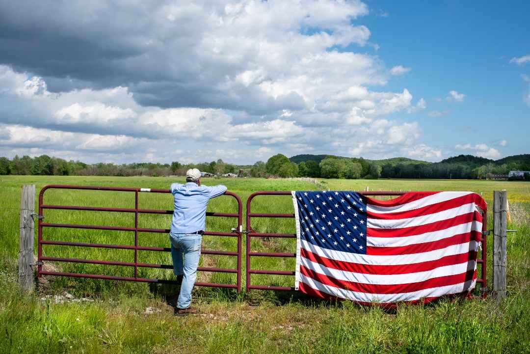 Famer leaning on fence looking over field with US flag hanging on gate.